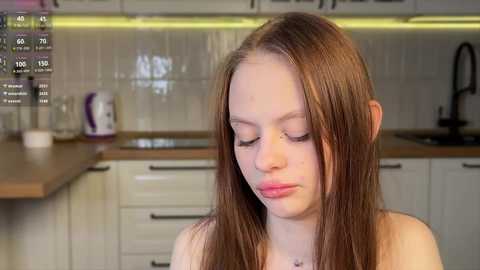 Media: A video of a young, fair-skinned woman with long brown hair, wearing a pink top, in a modern kitchen with white cabinets, glass jars, and a kettle on a wooden counter.