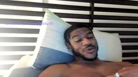 Media: Video of a shirtless Black man with a beard and short hair, lying on a bed with white and blue striped pillows and dark brown wooden blinds in the background.