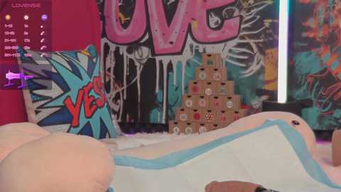 Media: Video of a colorful, vibrant room with a large \"LOVE\" mural, a plush bear, and a stack of boxes, featuring a video game interface on the left.