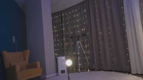 Media: Video of a dimly lit room with a gray curtain backdrop, a spotlight, a tripod, and an orange armchair.