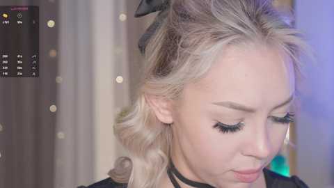 Media: Video of a young, fair-skinned woman with wavy, platinum blonde hair styled in soft waves, wearing black choker and black top, looking down. Background features soft, blurred lights and a black curtain.