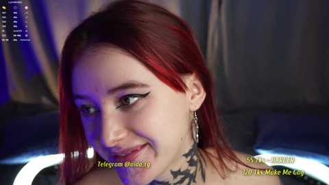 Media: Video of a young woman with bright red hair, light skin, and dark eyeliner, smiling, wearing a black, lace-up top, set in a dimly lit room with blue and purple lighting.