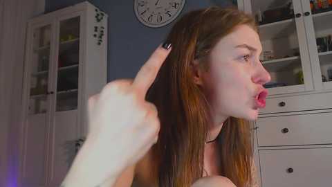 Media: Video of a young woman with long, straight red hair, wearing a black choker, making a middle finger gesture. Background features a white cabinet, clock, and shelves.