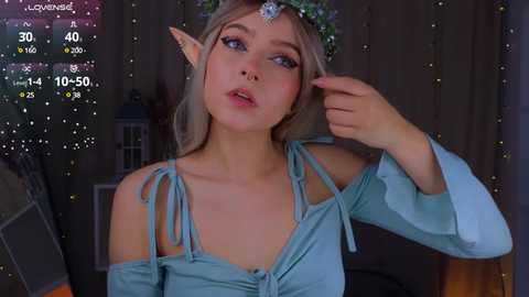 Media: A video of a young woman with light skin, pointed ears, and blonde hair, wearing a blue off-shoulder top with a leaf crown. The background features a dimly lit room with fairy lights and a digital weather display.