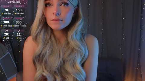 Media: Video of a young woman with long, wavy blonde hair, fair skin, and blue eyes, wearing a headband. She appears topless, in a dimly lit room with a virtual reality interface overlay.