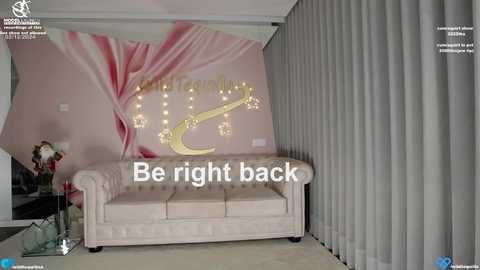 Media: Video of a pink-themed room with a tufted beige sofa, large floral mural, white curtains, and text overlay \"Be right back.\