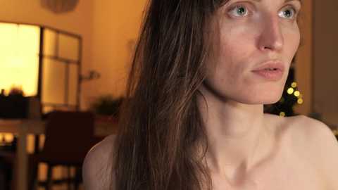 Media: A video of a young Caucasian woman with long, straight brown hair and fair skin, looking contemplative. Warm, yellowish lighting in a cozy room with a TV, chairs, and a Christmas tree in the background.