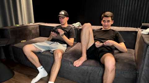 Media: Video of two young men, one in a baseball cap, the other in a cap, playing video games on a dark gray sectional sofa. They wear casual clothing and white socks.