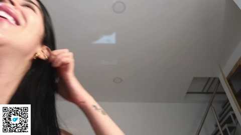 Media: Video of a smiling woman with long black hair, wearing gold hoop earrings, adjusting her earpiece in a modern, minimalist room with white walls and a mirror.