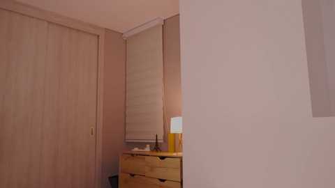 Media: A video of a minimalist bedroom corner featuring light wooden furniture, a small lamp, and a partially open closet door against pale beige walls.