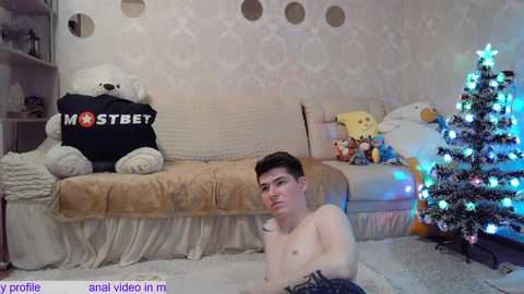 Media: A video of a shirtless young man sitting on the floor in front of a Christmas tree and a teddy bear bed, with a cozy, whimsical bedroom setting.