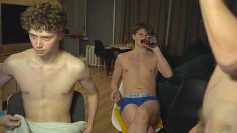 Media: Video of two shirtless, fair-skinned teenage boys in a dimly lit living room; one sips a drink, the other wears blue underwear, both in casual attire.