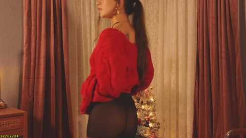 Media: A video of a woman in a red off-shoulder sweater and black tights, standing in a warmly lit room with a Christmas tree in the background.