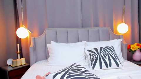 Media: Video of a modern bedroom with a gray upholstered headboard, white bedding, black zebra-patterned pillows, and two spherical pendant lights. Nightstand holds a candle, flowers, and a book.