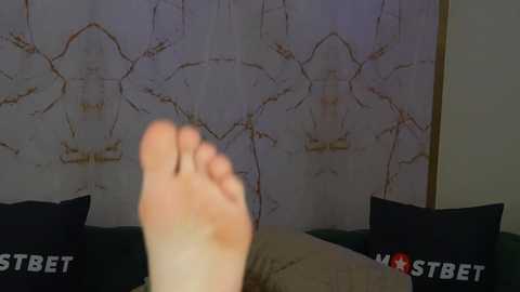 Media: Video of a person's foot in the foreground, with a dark curtain featuring white, abstract patterns in the background. The person is reclining on a bed with black pillows, and the \"MSTBET\" logo is visible in the lower right corner.