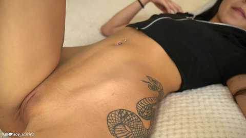 Media: Video of a light-skinned woman with a black snake tattoo on her right thigh, wearing a black crop top, lying on a beige couch.