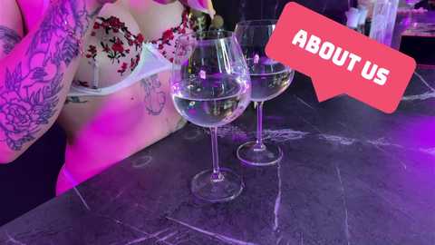 Media: Video of a tattooed woman in lingerie, holding two glasses of white wine, against a purple-lit marble countertop. Text reads, \"ABOUT US,\" in a red speech bubble.