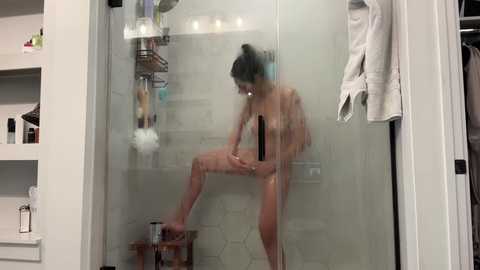 A video of a nude woman with dark hair in a bun, showering in a modern bathroom with white hexagonal tiles. She has a medium build and medium-sized breasts.