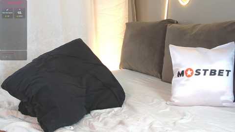 Video of a minimalist bedroom with white walls, a bed covered in white sheets, a black pillow, and a white pillow with \"Mostbet\" logo in black and orange text.