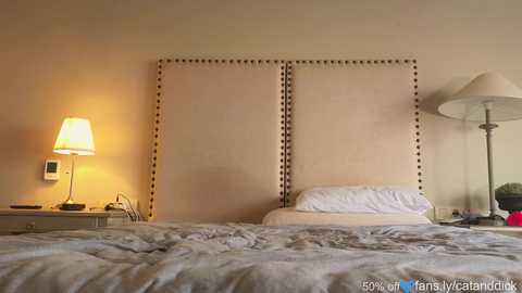 Media: Video of a cozy bedroom with a beige headboard, white bedding, a nightstand with a lamp, and a floor lamp, all in a neutral color scheme.