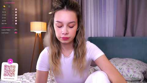 Media: Video of a young woman with light skin, long blonde hair, and a white t-shirt, sitting on a bed with a floral duvet, in a dimly lit bedroom.