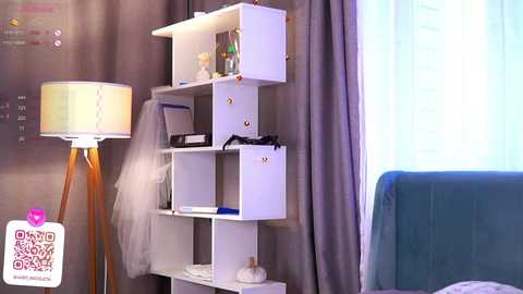 Media: Video of a modern, white shelving unit with drawers, adorned with a white tutu, a teal cushion, and a lamp with a beige lampshade, set against gray curtains.