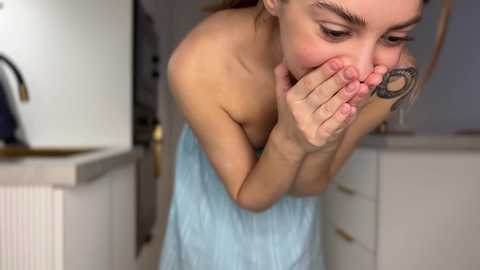 Media: Video of a young woman with fair skin, brown hair, and a tattoo on her hand, covering her mouth in a kitchen with light countertops and white cabinets.