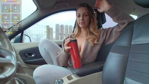 Media: Video of a blonde woman with long wavy hair, wearing a beige cardigan and light grey pants, sitting in a car, drinking from a red cup, with a city skyline visible through the window.