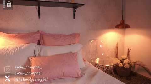 Media: A cozy bedroom with a warm, pink-toned atmosphere. The bed is neatly made with plush pink pillows, white sheets, and a headboard. A glass table with flowers and candles sits next to a modern chair, under a pendant light.