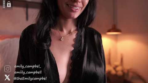 Video of a smiling, light-skinned woman with long black hair, wearing a black satin robe, in a cozy, dimly lit bedroom with a bedside lamp.