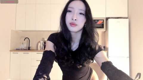 Media: A video of an Asian woman with long black hair, wearing a black lace top, standing in a modern kitchen with white cabinets and a fridge.