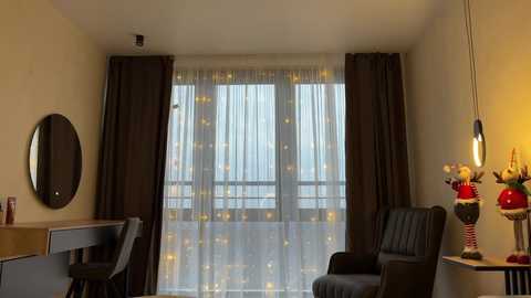 Media: Video of a modern, minimalist hotel room with a large window covered by sheer white curtains with gold patterns, dark brown curtains, and a black chair. A circular mirror hangs on the beige wall, and a wooden desk with a chair is visible on the left.