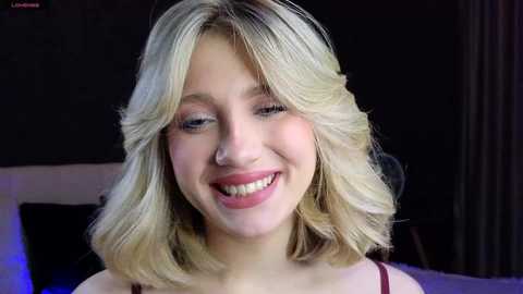 Media: Video of a young woman with blonde shoulder-length hair, fair skin, and red lipstick, smiling broadly. She wears a red top, set against a dark background with dim lighting.