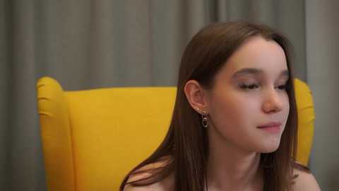 Video of a young woman with fair skin and long brown hair, wearing a yellow dress, seated in a bright yellow armchair. She has a serene expression, with closed eyes and a slight smile.