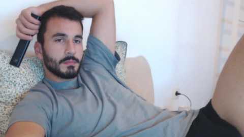 Media: Video of a bearded man with short dark hair, wearing a gray T-shirt and black shorts, relaxing on a patterned couch, holding a remote control.