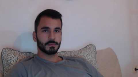 Media: A video of a young man with short black hair, a trimmed beard, and a gray shirt, lying on a beige couch with a patterned cushion against a plain white wall.