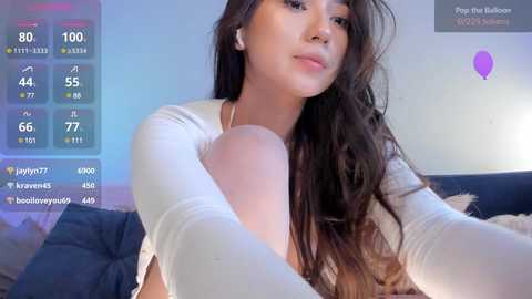 Media: Video of a young Asian woman with long, wavy brown hair, fair skin, and light makeup, wearing a white long-sleeve top. She's in a bedroom with a blue bedspread and a digital clock showing 100.