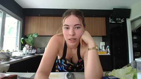 Media: Video of a young woman with light skin, brown hair, wearing a black and colorful floral apron, leaning on a kitchen counter in a modern, light-filled kitchen.