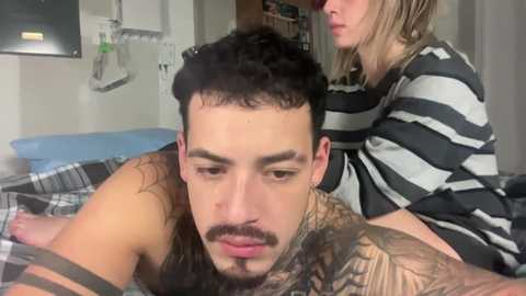 Media: Video of a man with a full beard and tattoos, shirtless, lying on a bed, with a woman in a striped shirt standing behind him.