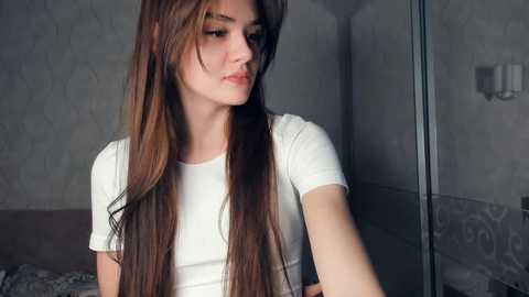 Media: Video of a young Caucasian woman with long, straight brown hair, wearing a white T-shirt, standing indoors next to a glass door, with a textured gray wall in the background.