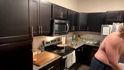 Media: Video of a modern, dark wood kitchen with stainless steel appliances, a microwave, and a white towel hanging. A topless person with light skin and short hair is cooking, facing away.