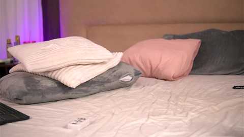 Video of a neatly made bed with beige sheets, two pillows in shades of gray and pink, and a TV remote on the right side.