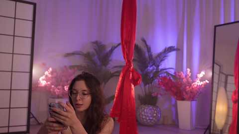 Media: Video of a young woman with long brown hair, wearing glasses and a red dress, taking a selfie in a dimly lit room with white curtains, purple lighting, and red and pink floral arrangements.