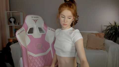 Media: Video of a young, fair-skinned woman with red hair in a ponytail, wearing a white crop top, standing beside a pink gaming chair in a cozy living room with beige walls and a white sofa.