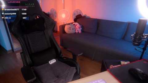 Media: A video of a cozy living room with a dark blue sectional sofa, a black gaming chair, and a pink stuffed animal. The room has a warm, dim lighting and a digital clock in the background.