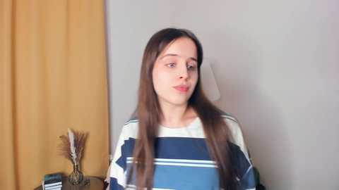 Media: Video of a young woman with long brown hair, wearing a striped shirt, standing in a room with beige walls, yellow curtains, and a small table with a lamp and decorative items.