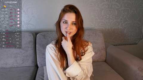 Media: Video of a young woman with long, wavy red hair, light skin, and a slender build, wearing a beige robe, sitting on a grey couch in a modern living room with a fancy wallpaper.