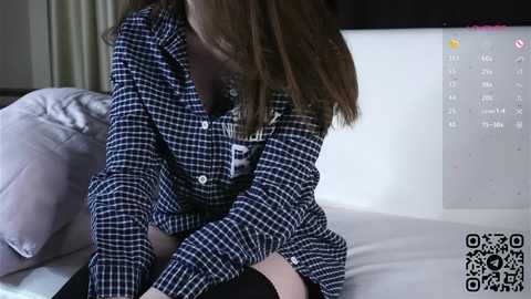 Media: Video of a young woman with long brown hair wearing a plaid shirt and black stockings, sitting on a bed with white sheets, in a modern bedroom.