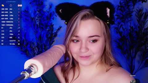 Media: Video of a young, fair-skinned woman with blonde hair, wearing black cat ears and smiling into a microphone, against a blue background with dark foliage.