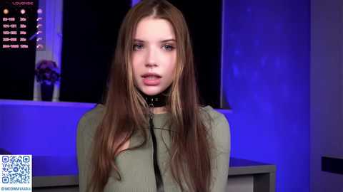 Video of a young Caucasian woman with long, straight brown hair, wearing a green sweater and black choker. Background features a dimly lit room with a TV displaying a news ticker and blue lighting.